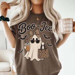 Peut inclure: Un t-shirt marron foncé avec un motif de fantôme portant des lunettes de soleil et un nœud à imprimé léopard, tenant un gobelet à café et un sac fourre-tout léopard. Le texte "Boo-Jee" est imprimé au-dessus, entouré de petites chauves-souris.