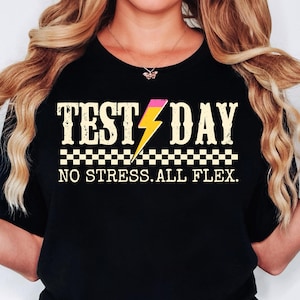 以下が含まれることがあります： 「テストデー」と雷と市松模様が描かれた白と黄色のグラフィックが施された黒いTシャツ。グラフィックの下のテキストは「ノーストレス。オールフレックス。」です
