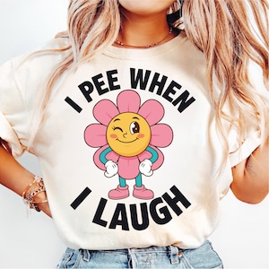 May include: Cream-colored t-shirt with the text "I PEE WHEN I LAUGH" in black. A cartoon flower with pink petals, a yellow center, and winking eyes is in the center of the shirt. The flower has blue arms and pink shoes.