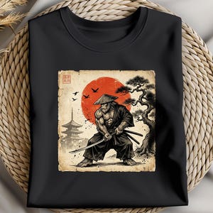 Peut inclure: T-shirt noir avec un graphisme de samouraï avec une épée, sur fond de soleil rouge et de paysage japonais. Le motif comprend une pagode, des oiseaux et un arbre stylisé. L'œuvre a une esthétique de papier vieilli vintage.