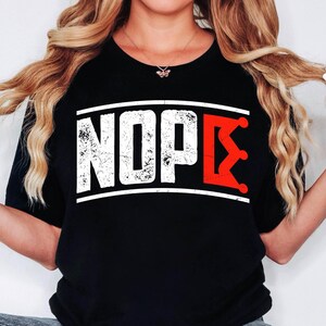Peut inclure: T-shirt noir avec le mot "NOPE" en blanc vieilli, encadré de blanc, et un motif de couronne rouge. Le t-shirt est de style décontracté, adapté au quotidien.