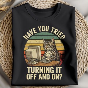 Peut inclure: T-shirt noir avec un chat devant un ordinateur et le texte "HAVE YOU TRIED TURNING IT OFF AND ON ?" Le design comprend un fond de coucher de soleil rétro. Le chat est brun et gris.