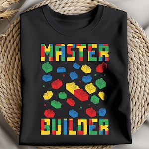 May include: Black t-shirt with the words "MASTER BUILDER" in colorful block letters. The shirt features a design of various colored toy building blocks scattered across the front, including red, yellow, blue, and green blocks.