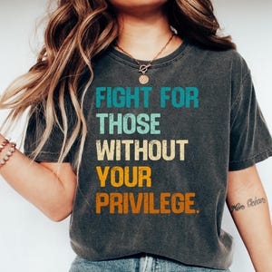 May include: A dark gray t-shirt with the text "FIGHT FOR THOSE WITHOUT YOUR PRIVILEGE." printed in colorful, vintage-style lettering.