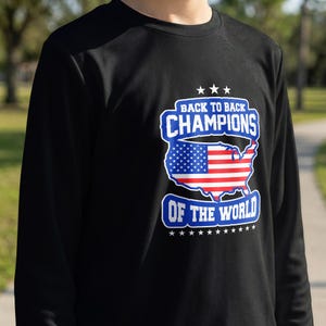 Peut inclure: T-shirt noir à manches longues avec un motif graphique. Le motif affiche le texte "BACK TO BACK CHAMPIONS OF THE WORLD" et une carte des États-Unis avec le drapeau américain.