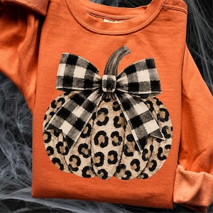 Peut inclure: Un sweat-shirt orange à manches longues arborant un motif central de citrouille avec un imprimé léopard beige et marron. La citrouille est surmontée d'un grand nœud décoratif à motif de carreaux vichy noirs et blancs.