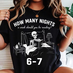 Op de afbeelding: Zwart T-shirt met een witte afbeelding van een skelet dat in bed leest, met een nachtkastje, lamp en tekst: "HOW MANY NIGHTS a week should you be reading?" en "6-7".