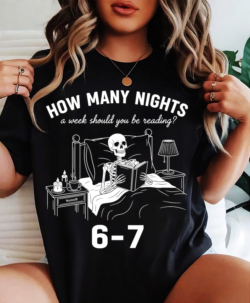 Op de afbeelding: Zwart T-shirt met een witte afbeelding van een skelet dat in bed leest, met een nachtkastje, lamp en tekst: "HOW MANY NIGHTS a week should you be reading?" en "6-7".