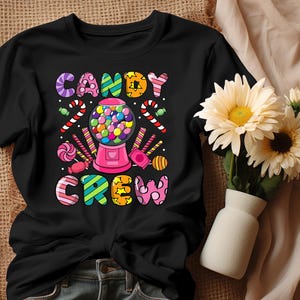 Peut inclure: T-shirt noir avec l'inscription colorée "CANDY CREW". Le motif comprend une machine à mâcher, des cannes en sucre et divers bonbons. Le t-shirt est en matière souple, idéal pour un look décontracté.