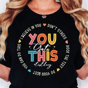 Pode incluir: Camiseta preta com um gráfico colorido que diz "You Got This" com corações e o texto "I believe in you, don't stress, rock the test, do your best, you can do this #testday".
