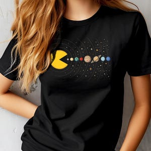 May include: Black t-shirt featuring a yellow Pac-Man character consuming the planets of the solar system. The planets are depicted in their respective colors and positions within the solar system. The design includes stars and curved lines.