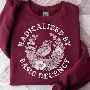 Peut inclure: Un sweat-shirt bordeaux avec le texte "RADICALIZED BY BASIC DECENCY" dans un design circulaire. Le motif présente une illustration détaillée d'un oiseau perché parmi des fleurs et du feuillage, le tout en blanc.