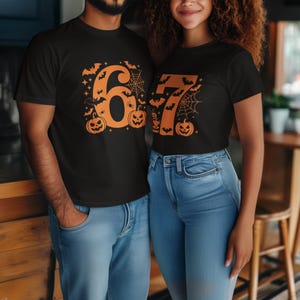 May include: Black t-shirts with orange Halloween-themed designs. One shirt features the number "6" with bats, spiders, and pumpkins. The other shirt displays the number "7" with similar Halloween elements. Both shirts are paired with blue jeans.