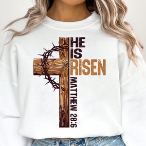 May include: White sweatshirt with a brown wooden cross and a crown of thorns. The text "HE IS RISEN MATTHEW 28:6" is printed on the sweatshirt.