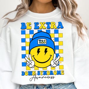 May include: White sweatshirt with a blue and yellow checkered background. A yellow smiley face with lightning bolts for eyes wears a blue beanie with the letters "T21" on it. The text "BE EXTRA DOWN SYNDROME Awareness" is printed on the sweatshirt.