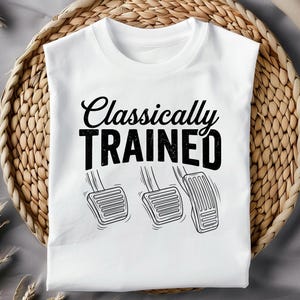 May include: White t-shirt with the text "Classically TRAINED" in black, above a graphic of three car pedals. The shirt is displayed on a woven wicker tray.