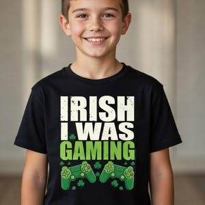Peut inclure: Un t-shirt noir avec l'inscription "IRISH I WAS GAMING" en blanc et vert. Le motif comprend des trèfles verts et deux manettes de jeu vertes. Le t-shirt est porté par un enfant souriant.
