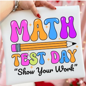 以下が含まれることがあります： 「MATH TEST DAY」の文字がバブルレターで書かれた白い正方形のサイン、黄色の鉛筆、そして「Show Your Work」というフレーズ。文字はピンク、オレンジ、青、緑、紫です。