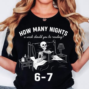 Op de afbeelding: Zwart t-shirt met een witte afbeelding van een skelet dat in bed leest, met een nachtkastje, een lamp en kaarsen. De tekst luidt "HOW MANY NIGHTS a week should you be reading?" en "6-7".