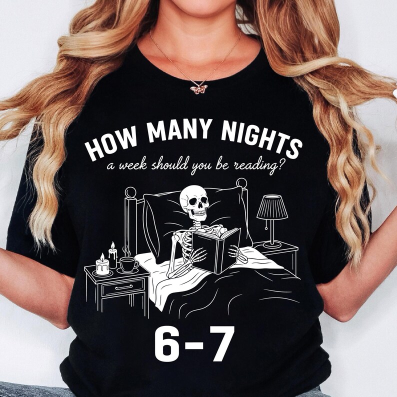 Op de afbeelding: Zwart t-shirt met een witte afbeelding van een skelet dat in bed leest, met een nachtkastje, een lamp en kaarsen. De tekst luidt "HOW MANY NIGHTS a week should you be reading?" en "6-7".