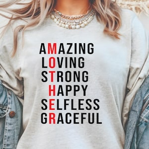 Może przedstawiać: Jasnoszara koszulka z napisem "MOTHER" zapisanym pionowo czarnymi i czerwonymi literami. Słowa "AMAZING, LOVING, STRONG, HAPPY, SELFLESS, GRACEFUL" są ułożone pionowo, aby opisać słowo "MOTHER".