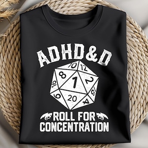 以下が含まれることがあります： 黒いTシャツ。白の20面ダイスの上に「ADHD & D」の文字、数字の1が大きく表示されています。「ROLL FOR CONCENTRATION」のフレーズがダイスの下にあります。