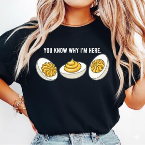 Peut inclure: Sweat-shirt noir avec l'inscription "YOU KNOW WHY I'M HERE." et un motif de trois œufs farcis. Les œufs sont blancs avec une garniture jaune.