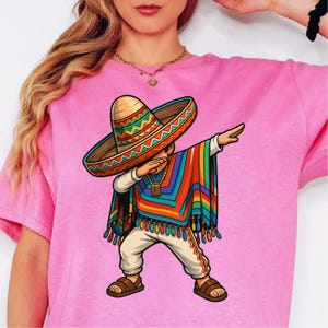 Peut inclure: T-shirt rose avec une illustration de dessin animé d'une personne faisant un dab, portant un sombrero, un poncho coloré et des vêtements mexicains traditionnels. Le design comprend des couleurs vives et des détails complexes.