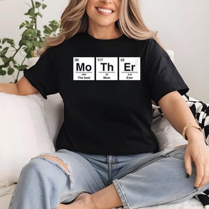 Peut inclure: T-shirt noir avec le mot "Mother" épelé à l'aide du tableau périodique des éléments. Le t-shirt présente les éléments Mo, Th et Er, avec les mots "The Best", "Mom" et "Ever" sous chaque symbole d'élément. La personne porte un jean bleu.