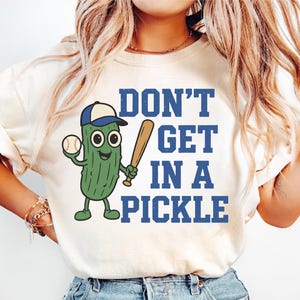 May include: Cream-colored t-shirt with a cartoon pickle character wearing a baseball cap and holding a baseball and bat. The text "DON'T GET IN A PICKLE" is printed in blue block letters.