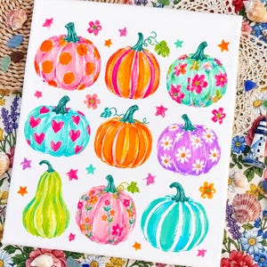 Peut inclure: Une toile blanche présentant une grille de neuf citrouilles peintes avec des couleurs et des motifs variés, incluant des pois, des rayures, des cœurs et des fleurs, agrémentée de petites étoiles et fleurs peintes sur toute la surface.