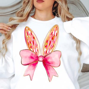 Peut inclure: Sweat-shirt blanc avec un nœud rose et des oreilles de lapin avec des accents de paillettes dorées. Le nœud est grand et a un effet aquarelle.
