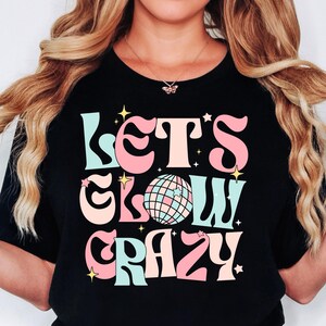 Peut inclure: T-shirt noir avec un graphisme rétro qui dit "Let's Glow Crazy" en rose, bleu et jaune. Un ballon disco est présenté au centre du graphisme.
