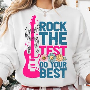 May include: White sweatshirt with a vibrant graphic. The design features a pink electric guitar, musical notes, and the text "ROCK THE TEST DO YOUR BEST" in blue and pink. Smiling flower graphics add a cheerful touch.