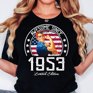 Può includere: T-shirt nero con grafica di Rosie the Riveter e la scritta "Awesome Since 1953 Limited Edition." Il design include la bandiera americana e stelle. La maglietta ha un girocollo.