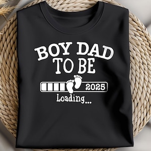 Peut inclure: T-shirt noir avec l'inscription blanche "BOY DAD TO BE" au-dessus d'une barre de chargement avec des empreintes de pieds de bébé et l'année "2025". Le mot "Loading..." est en dessous.