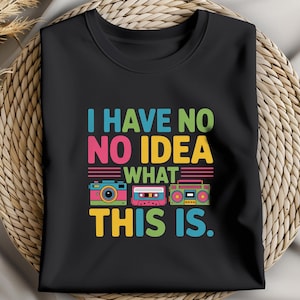 Puede incluir: Camiseta negra con la frase "I HAVE NO IDEA WHAT THIS IS". El diseño incluye ilustraciones coloridas de una cámara, una cinta de cassette y un radiocasete. La camiseta se muestra sobre una superficie tejida.