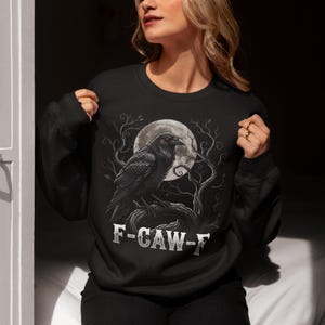 May include: Black sweatshirt with a raven illustration set against a full moon and bare branches. The design includes the text "F-CAW-F". The raven is depicted with red eyes. A gothic-themed design.