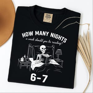 Op de afbeelding: Zwart T-shirt met een witte afbeelding van een skelet dat in bed leest, met de tekst "HOW MANY NIGHTS a week should you be reading?" en "6-7". Het shirt heeft een ronde hals en een klein label bij de halslijn.