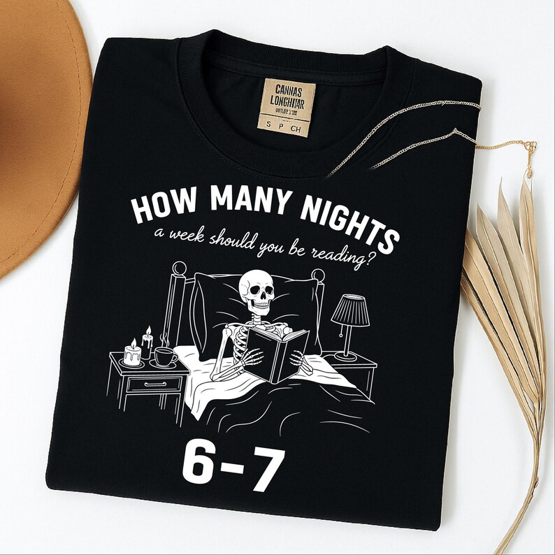 Op de afbeelding: Zwart T-shirt met een witte afbeelding van een skelet dat in bed leest, met de tekst "HOW MANY NIGHTS a week should you be reading?" en "6-7". Het shirt heeft een ronde hals en een klein label bij de halslijn.
