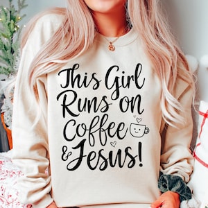 Puede incluir: Una sudadera beige con el texto "This Girl Runs on Coffee & Jesus!" en negro. Una pequeña taza de café con una carita sonriente está al lado del texto.