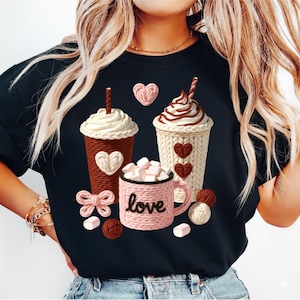 Peut inclure: Sweat-shirt noir avec un motif de trois boissons chaudes avec de la crème fouettée et des cœurs. Une tasse rose avec le mot "love" et des guimauves est également représentée. Des boules de fil et un nœud complètent le design.