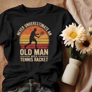 Peut inclure: T-shirt noir avec un motif sur le thème du tennis. Le design présente une silhouette de joueur de tennis avec le texte "NEVER UNDERESTIMATE AN OLD MAN WITH A TENNIS RACKET" dans un style coucher de soleil rétro. Un vase de fleurs est en arrière-plan.