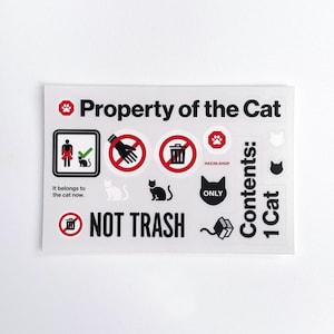 May include: A clear sticker sheet with various cat-themed graphics and text. The text includes "Property of the Cat," "Not Trash," and "It belongs to the cat now." The sheet features cat silhouettes, paw prints, and prohibition symbols.