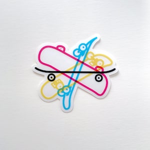 May include: A sticker featuring a stylized design of three overlapping skateboards in pink, yellow, and blue. The central skateboard is white with a black line, and the wheels are black circles. The sticker is on a white background.