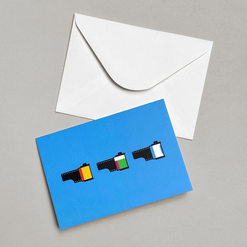May include: A blue greeting card with three stylized film roll illustrations in yellow, green, and white. A white envelope is in the background. The card is on a gray surface.