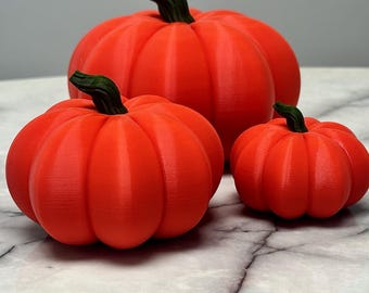Puede incluir: Tres calabazas naranjas con tallos verdes de diferentes tamaños sobre una superficie de mármol. Las calabazas tienen una textura acanalada, ideales para decoraciones de otoño o Halloween.