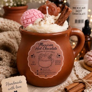 May include: A brown ceramic mug filled with a soy wax candle designed to look like Mexican hot chocolate. The candle features white wax whipped cream, a pink concha, a brown bear-shaped piece, and a cinnamon stick. The label reads Mexican Hot Chocolate.