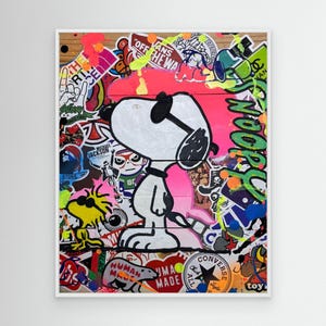 May include: A framed artwork featuring Snoopy from Peanuts, surrounded by colorful stickers and graffiti. The artwork has a pink and wooden background, with the word "NOOR" in green. The stickers include text like "Vans", "Human Made", and "Converse".