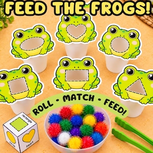 May include: A set of six white cups with green frog cutouts featuring various shapes including a heart, circle, square, rectangle, and triangle. Includes a white die with shape labels, a bowl of colorful pom-poms, and green plastic tongs.